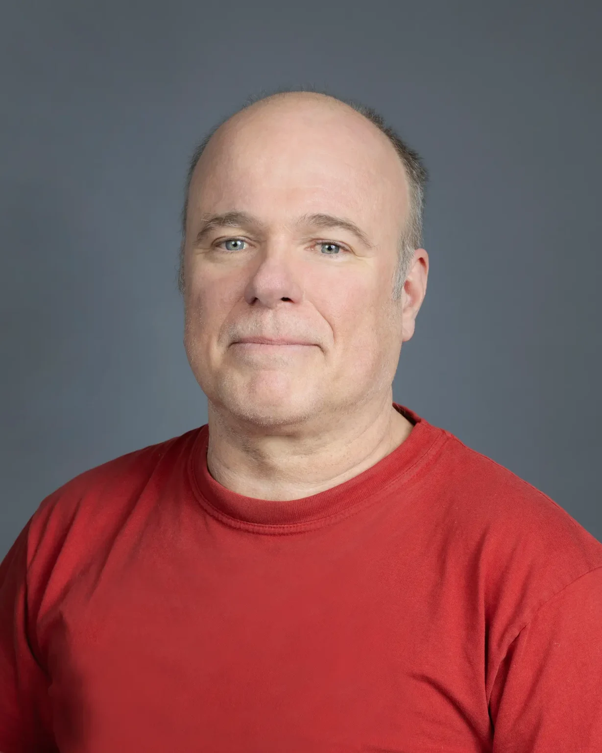 Close-up portrait of a man with light eyes and a friendly smile, dressed in a casual red t-shirt, centered against a solid grey backdrop